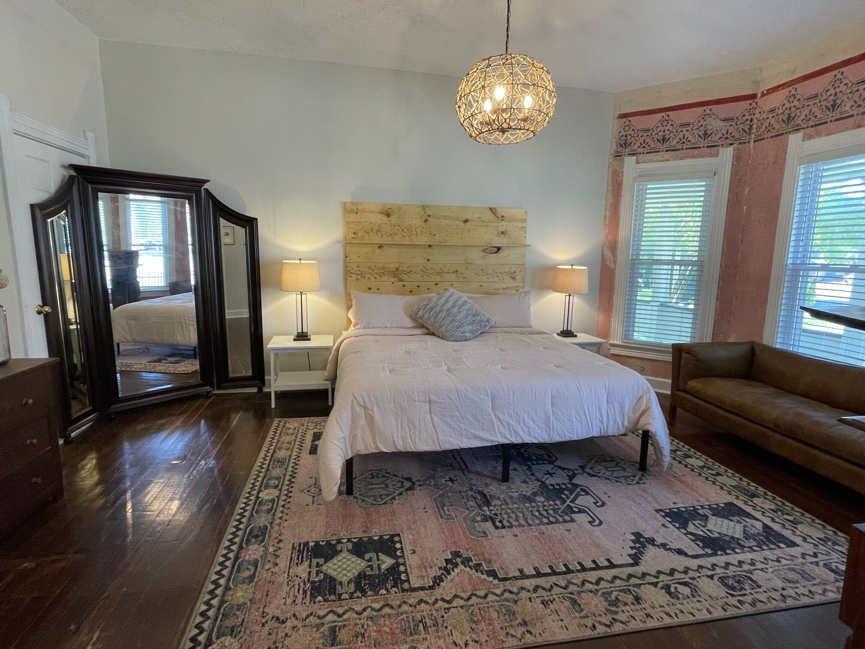 Une chambre à coucher avec une tête de lit en bois, un grand lit avec une literie blanche, deux lampes de chevet, un luminaire suspendu, un tapis à motifs et un fauteuil en cuir marron.