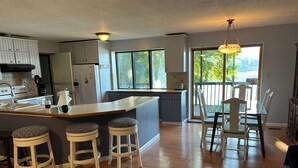 A modern kitchen with a central island, bar stools, a dining table with chairs, and a view through large windows. - 4 Bedroom Lake House - Northern Michigan (Central Lake)