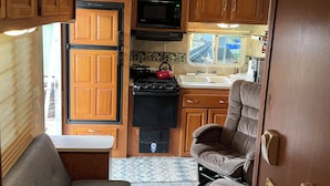 A compact RV kitchen with wooden cabinets, a microwave, stove, and a sink. There is a gray couch and a window providing natural light. - Cozy RV in a vacation community on Lake Hartwell (Westminster)