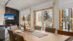 A dining area with a stone fireplace, a large window, and a view of trees. - RMR: Teton Pines 3443 Townhome in Wilson, WY (Wilson)