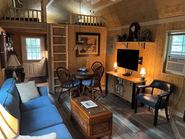 A cozy living room with a sofa, a dining table, and chairs. There's a TV, a bookshelf, and a fireplace.