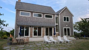 Ein zweistöckiges Haus mit überdachter Terrasse, Sitzgelegenheiten im Freien und einem gepflasterten Weg.