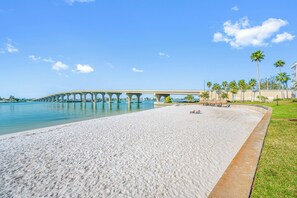 Uma praia com um píer de concreto que se estende para a água, uma área gramada com palmeiras e um céu azul límpido.