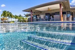 Ein Schwimmbecken mit klarem Wasser und Stufen zum hineinsteigen, umgeben von einer überdachten Außenbereich mit Stühlen und Tischen.