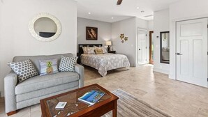 Een moderne woonkamer met een bank, een salontafel, een bed met hoofdbord, een plafondventilator en een ronde spiegel aan de muur.