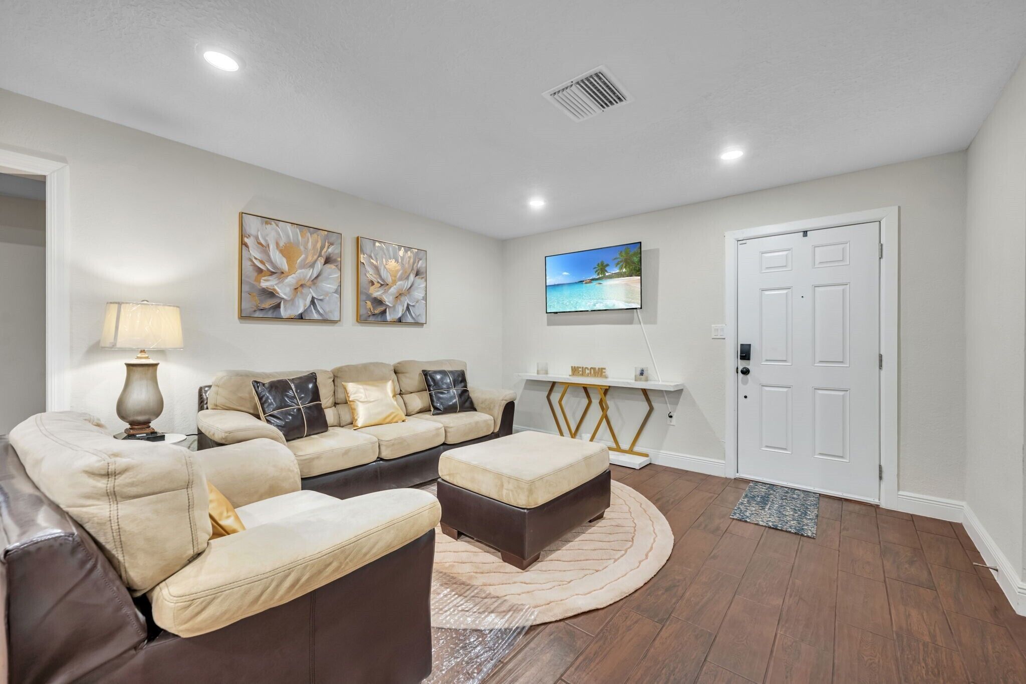 A living room with a beige sectional sofa, a small ottoman, a lamp, and wall art.
