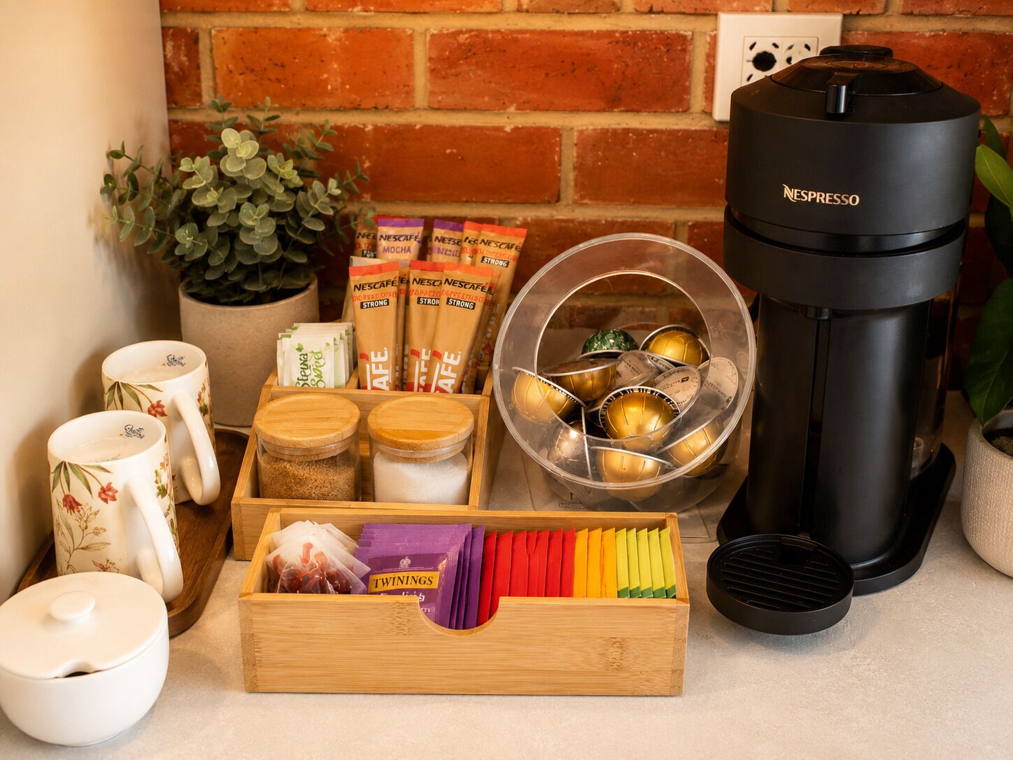 Una cocina con pared de ladrillo, una cafetera Nespresso, una tetera, una planta y varios objetos relacionados con el café.