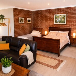 Un dormitorio con pared de ladrillo, dos camas, cabeceros de madera, un sofá de cuero y una pequeña mesa con una planta.