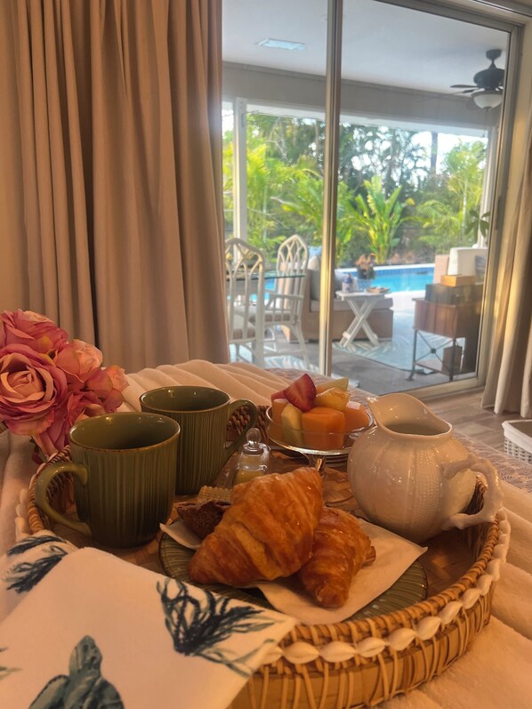 Ett frukostbricka med croissanter, en fruktskål och en kanna kaffe, placerat på ett bord med utsikt över en pool och grönområden.