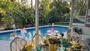 Ein Essbereich im Freien mit einem Glastisch, gedeckt für eine Mahlzeit, umgeben von Korbstühlen, in der Nähe eines Swimmingpools mit dekorativen Flamingoschwimmern.