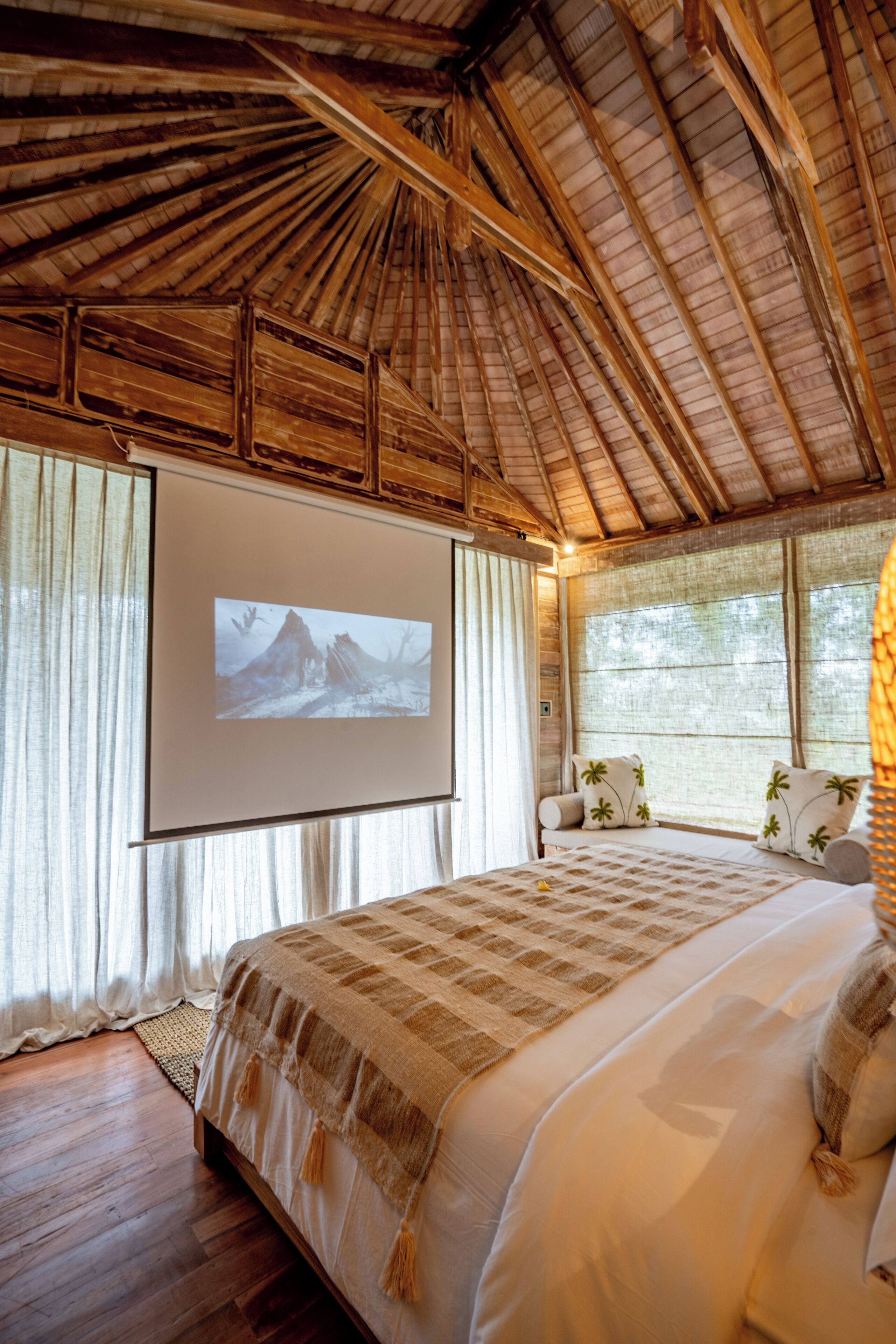 A spacious bedroom with a large bed, a projector screen on the wall, and a view of the outdoors through large windows.