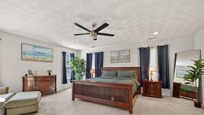 Quarto com cama de madeira, ventoinha no teto, mesa de cabeceira, cômoda e uma janela grande com cortinas.