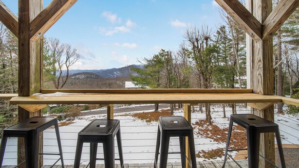 Eine Holzterrasse mit einem Holztisch und vier schwarzen Hocker. Die Terrasse bietet Blick auf eine verschneite Landschaft mit Bäumen und Bergen im Hintergrund.
