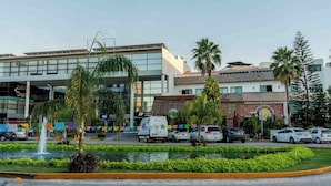 Un edificio moderno con un amplio muro de cristal, un estacionamiento con varios coches y una pequeña fuente al frente.