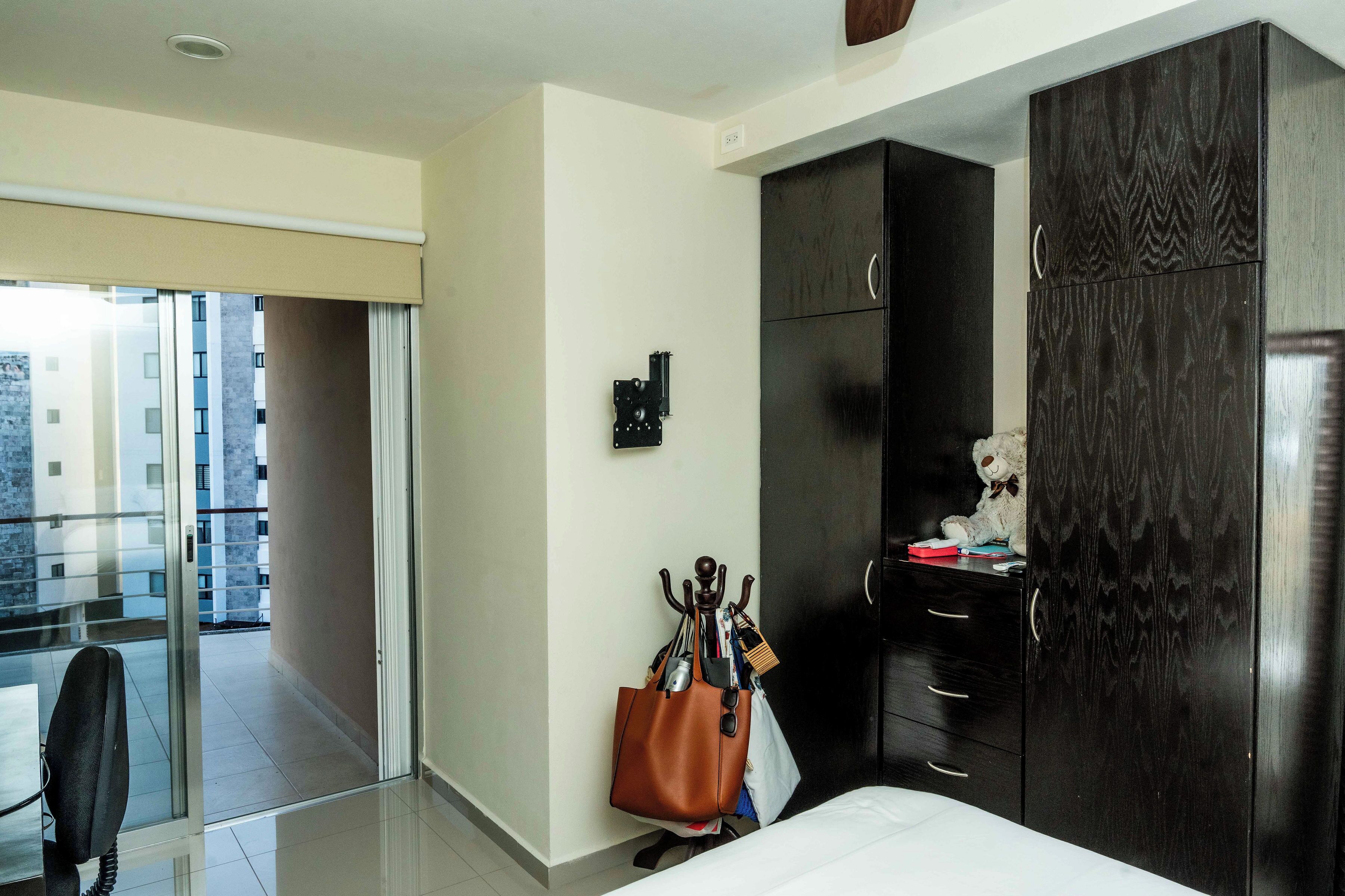 A hotel room with a bed, a wardrobe, a desk with a chair, a coat rack with bags, and a view of the outside balcony.