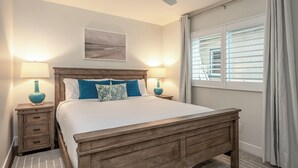 Ein Schlafzimmer mit einem hölzernen Bett, zwei Nachttischen mit Lampen, einem Deckenventilator, einem Fenster mit Jalousien und einem Wandbild im Strandhausstil.