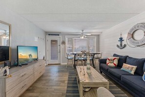 Una sala de estar con televisor, muebles de madera, un sofá, una mesa de comedor con sillas y un ventilador de techo.