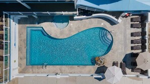 En vy från ovan av en swimmingpool med solstolar och parasoller, omgiven av en terrass med kakelplattor.