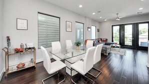 Una sala moderna con una mesa de comedor de vidrio, sillas blancas y un sofá. Hay una mesa de centro, una estantería y una planta sobre la mesa.