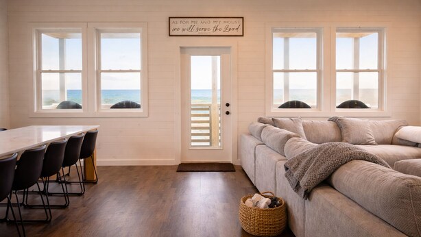 Une pièce avec un plafond en bois, une porte blanche donnant sur la plage, un canapé beige et une table à manger avec des chaises.