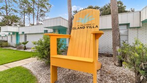 Una poltrona Adirondack gialla con il logo "The Iconic Island Villa", immersa in un giardino curato e affiancata da un edificio dalle rifiniture bianche e verdi.