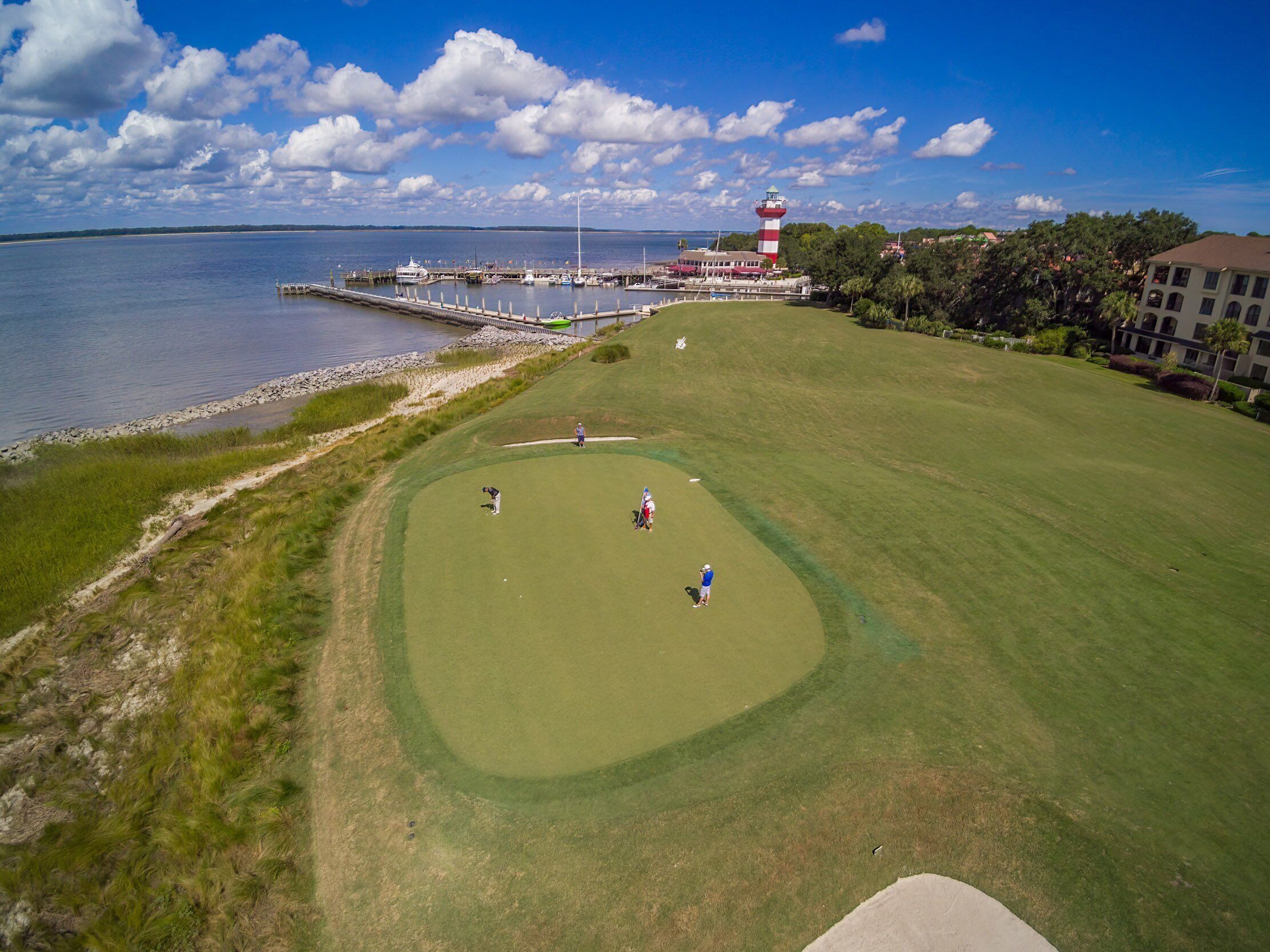 A golf course with a lighthouse in the background, a dock with boats, and a building with a red and white striped roof.
