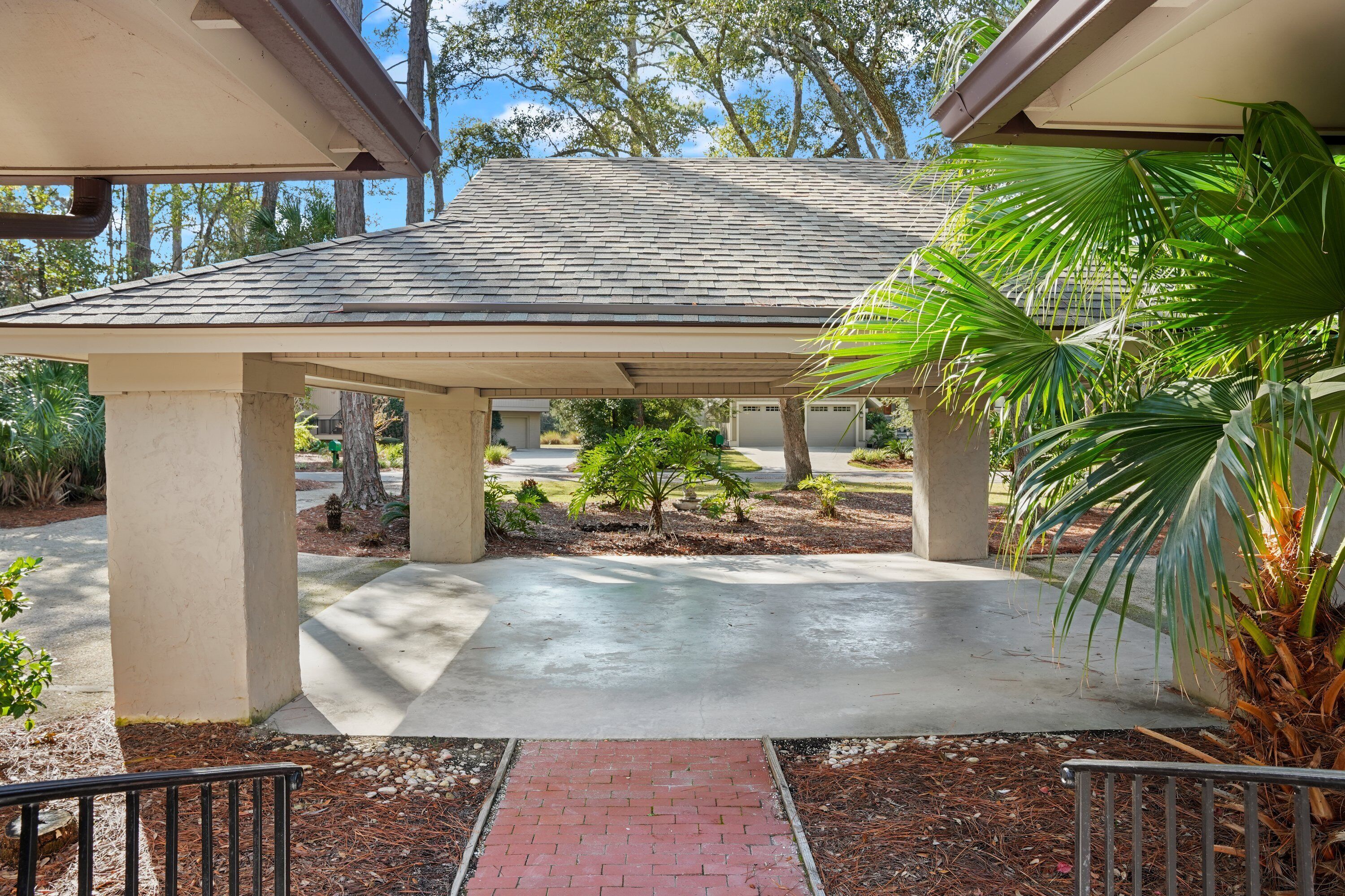 A covered outdoor area with a concrete floor, palm trees, and a brick pathway leading to a building entrance.