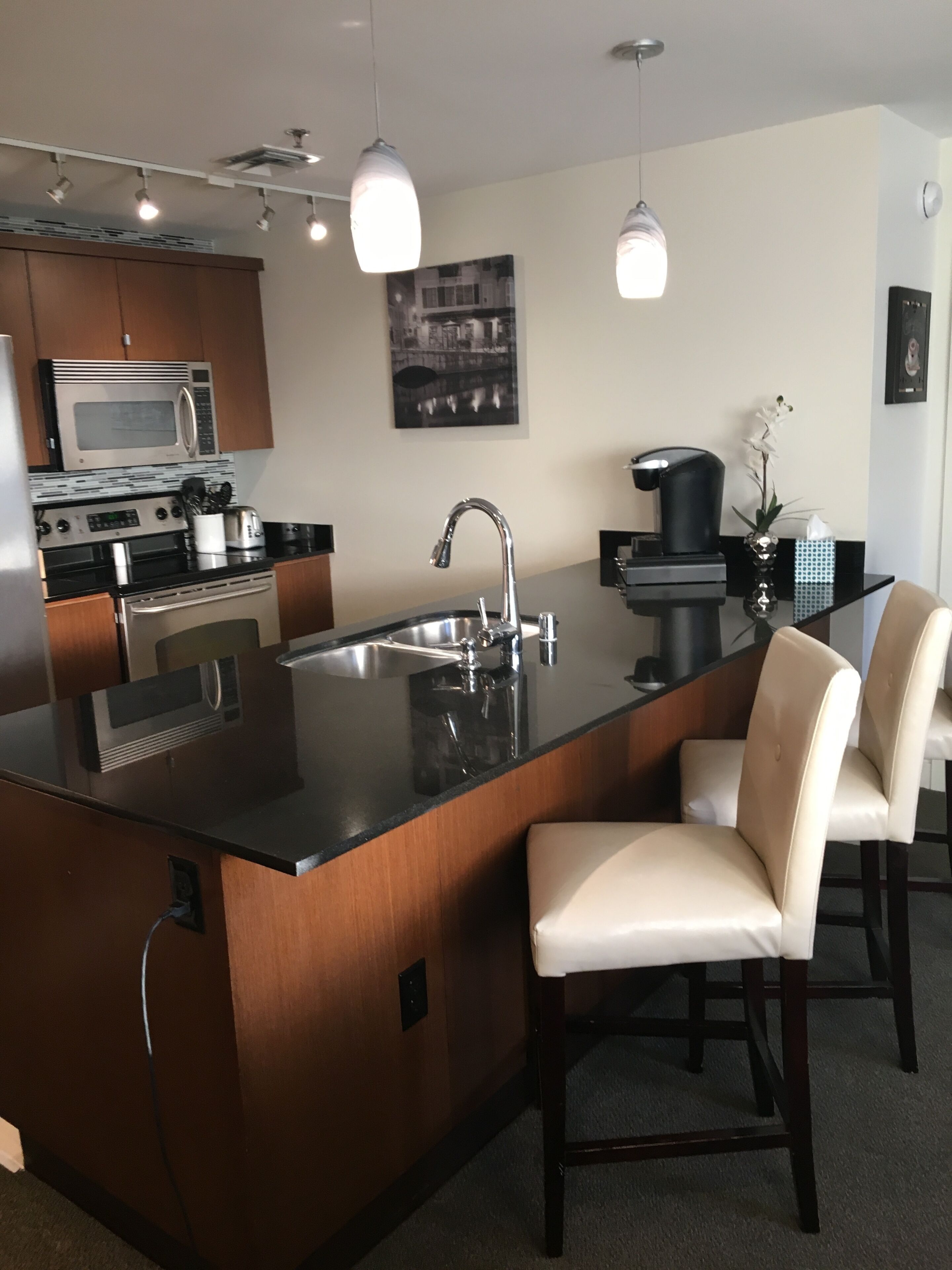 Una cocina moderna con una isla central, electrodomésticos de acero inoxidable, iluminación colgante y una cafetera de color negro.