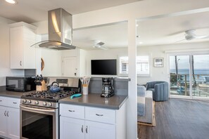 A modern kitchen with a gas stove, microwave, and coffee maker. There is a large window with a view of the ocean. - Steps to the Shore – Top Floor 3BR Ocean View -  #4 (Oceanside)