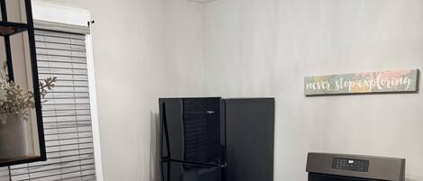 Una cocina compacta con un refrigerador, estufa y microondas negros. Hay un pequeño bote de basura y una planta en maceta en el alféizar.