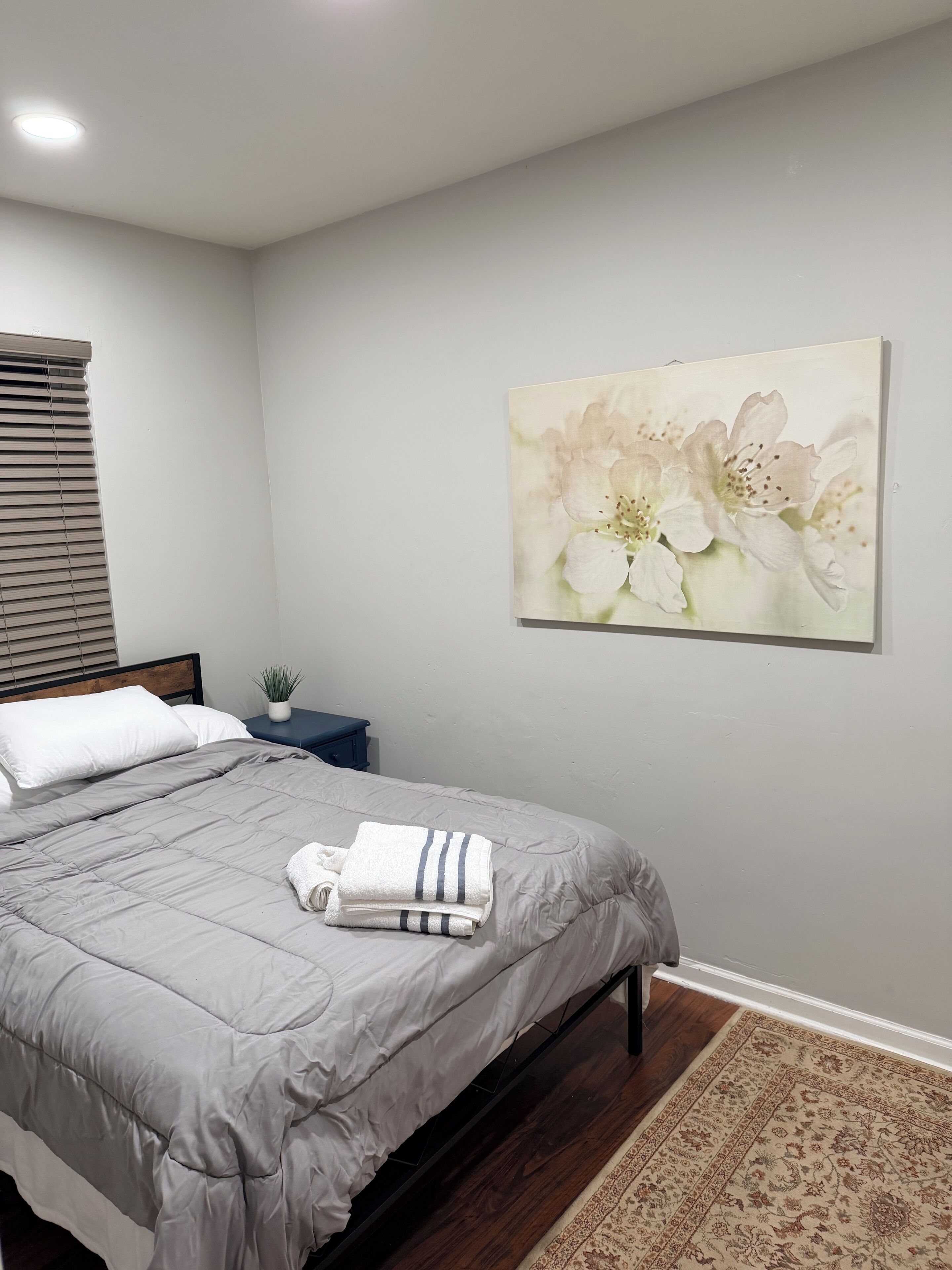 A neatly made bed with a grey duvet, a small bedside table with a plant, and a large floral painting on the wall.