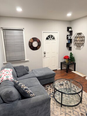 Una sala de estar con un sofá seccional gris, una mesa de centro redonda de vidrio, una puerta blanca, un reloj de pared y una estantería decorativa.
