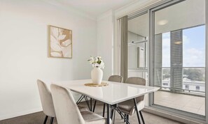 A modern room with a white table, four chairs, a vase with flowers, a framed artwork, and a large window with a view of buildings. - KozyGuru | Granville | Contemporary Living Retreat (Granville)