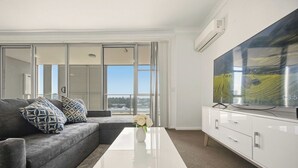 Ein modernes Wohnzimmer mit einem grauen Ecksofa, gemusterten Kissen, einem weißen Couchtisch, einem Flachbildfernseher und einem großen Fenster mit Blick auf die Stadt.