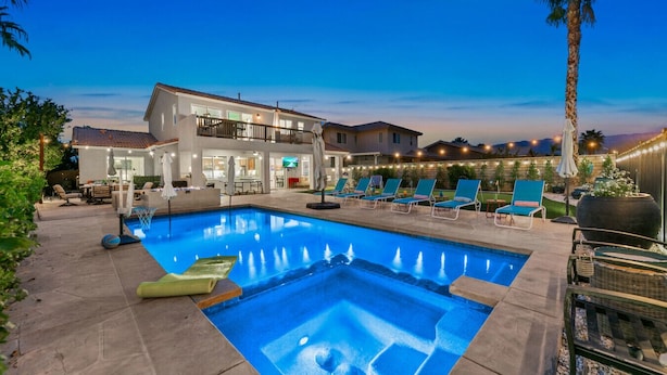 A swimming pool with lounge chairs, a covered patio area, and a two-story building with outdoor lighting. - Golf, Lake & Mountain Views, Pool, Spa, BBQ, Games (Indio)