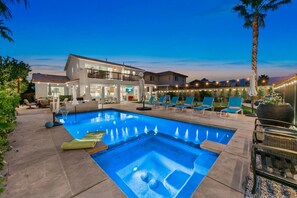 A swimming pool with lounge chairs, a covered patio area, and a two-story building with outdoor lighting.