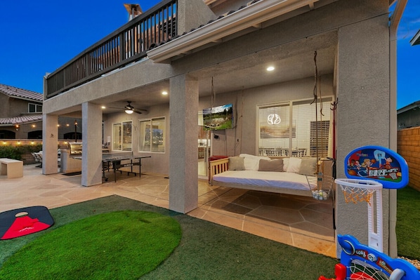 A two-story house with a balcony, a patio with a table and chairs, a swing, and a basketball hoop.