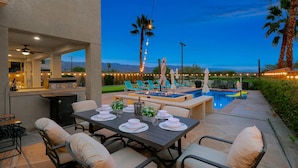 An outdoor patio with a dining table set for four, a grill, and a pool area with lounge chairs and umbrellas. - Golf, Lake & Mountain Views, Pool, Spa, BBQ, Games (Indio)