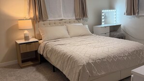 Ein Schlafzimmer mit Bett, Nachttisch, Lampe, Schminktisch mit Beleuchtung und Vorhängen.