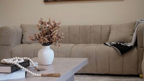 Ein Wohnzimmer mit einem beigefarbenen Sofa, einer weißen Vase mit Trockenblumen, einem Holztisch und einem gerahmten abstrakten Gemälde an der Wand.