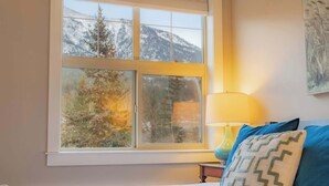 Ein Schlafzimmer mit Bett, Nachttisch, Lampe und einem Fenster mit Blick auf Berge und Bäume.