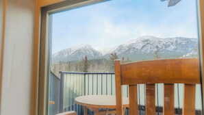 Ein Balkon mit Holzstuhl und Tisch, mit Blick auf eine gebirgige Landschaft mit schneebedeckten Gipfeln und bewaldetem Gebiet.