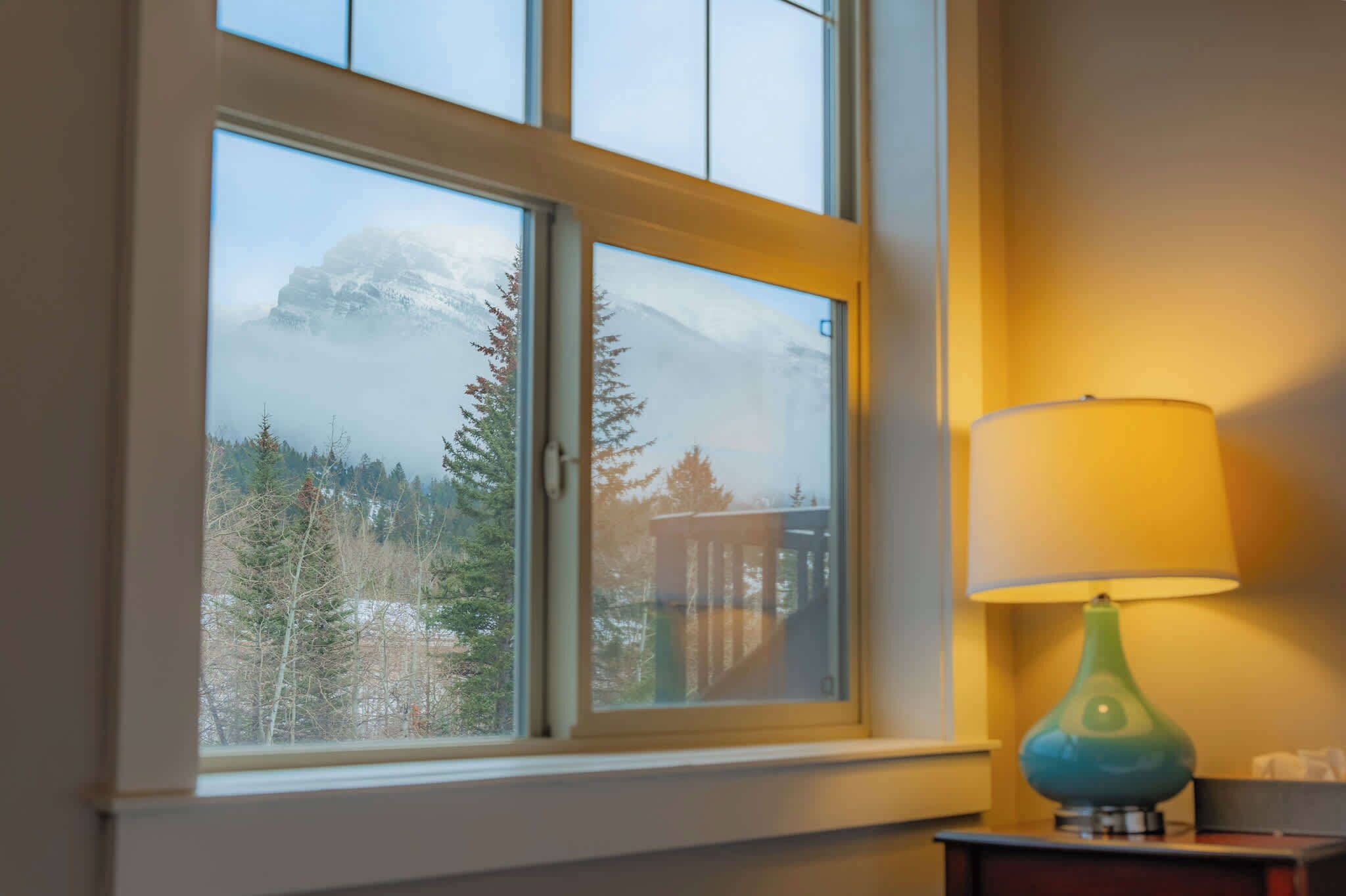 Una ventana con vistas a montañas nevadas y árboles, una lámpara en una mesita de noche y una mesita de noche de madera.