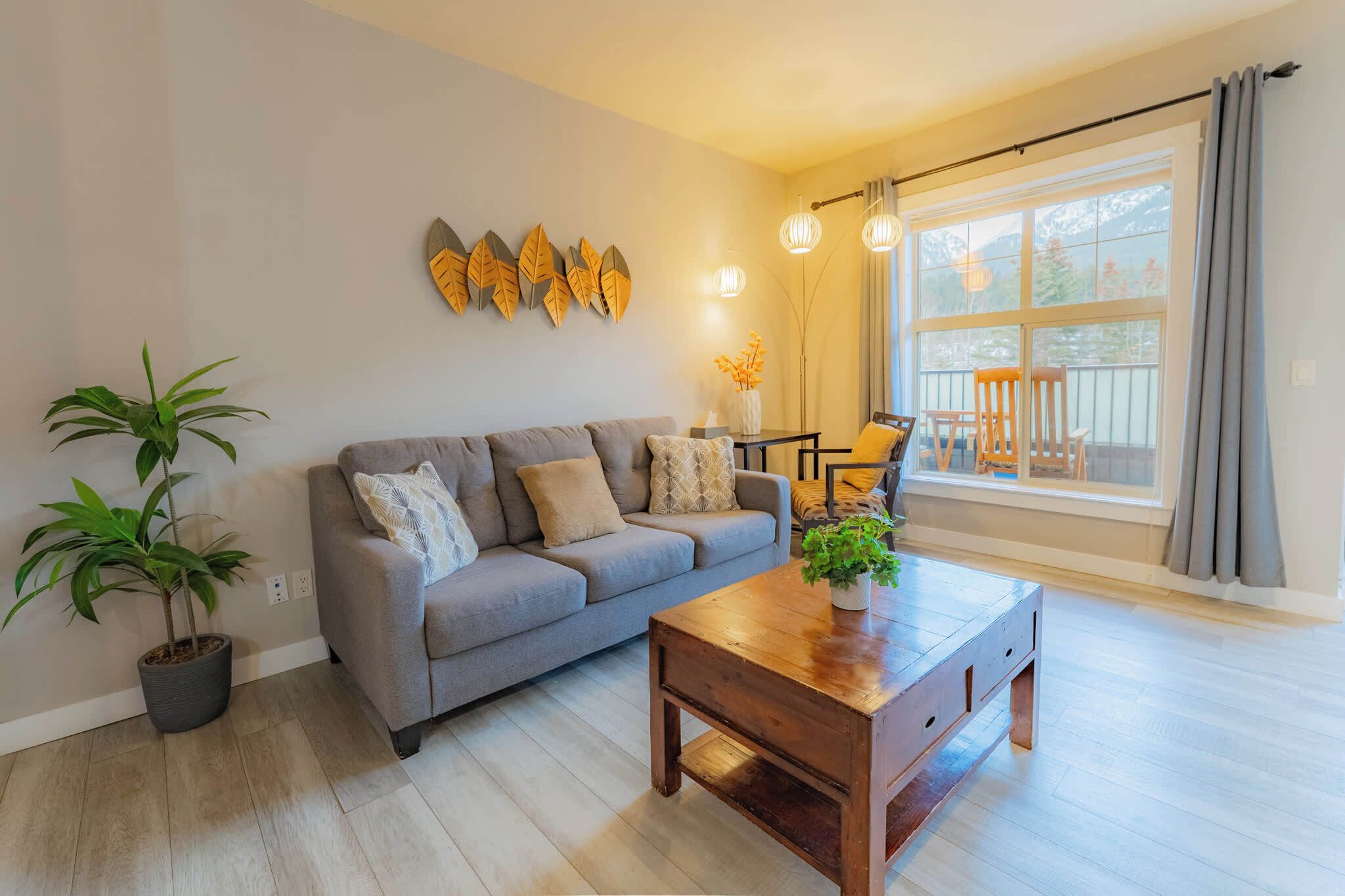 A living room with a grey sofa, a wooden coffee table, a potted plant, and a large window with curtains.