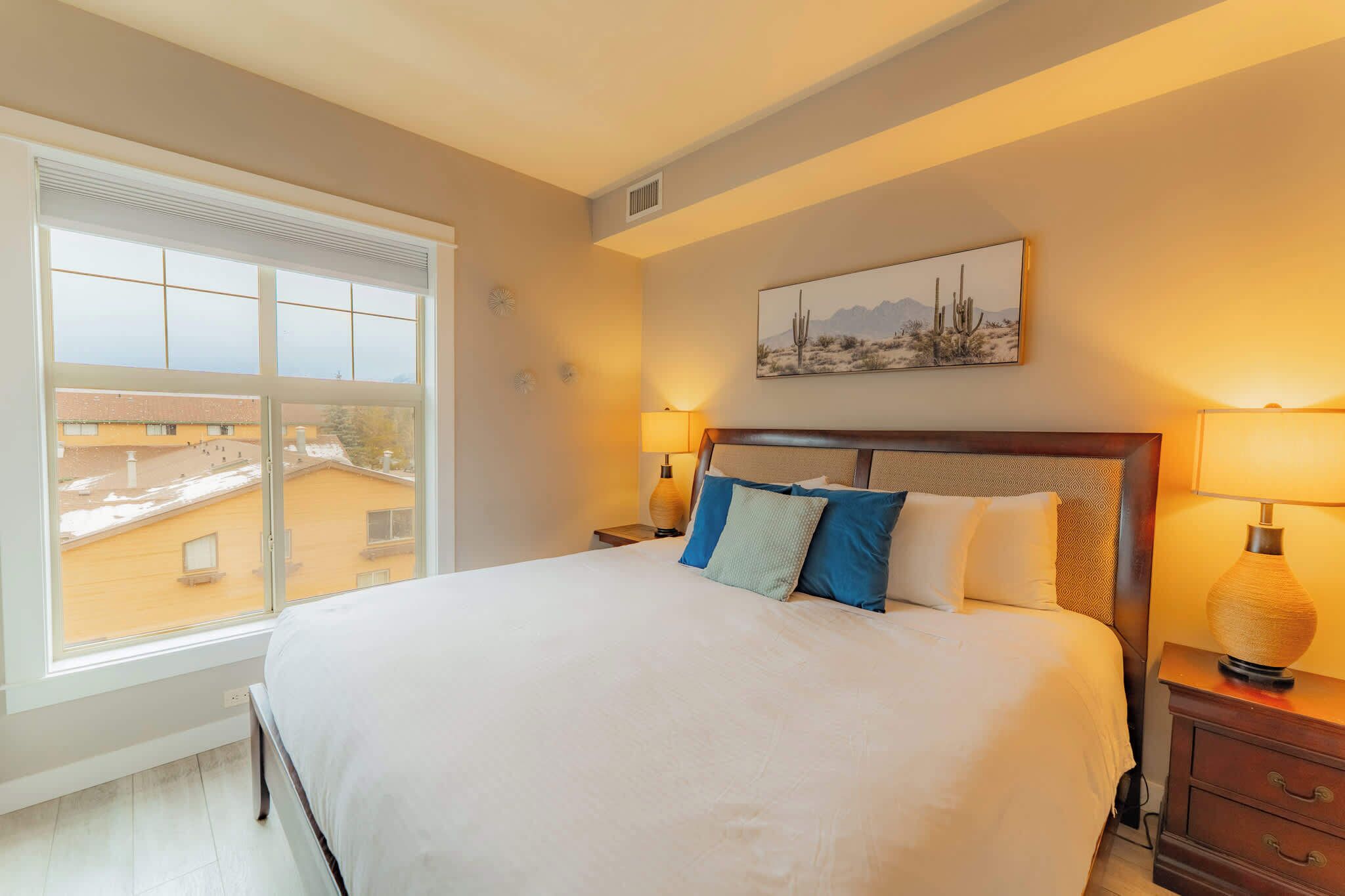 A neatly made bed with a headboard, two bedside lamps, and a window with a view of buildings.