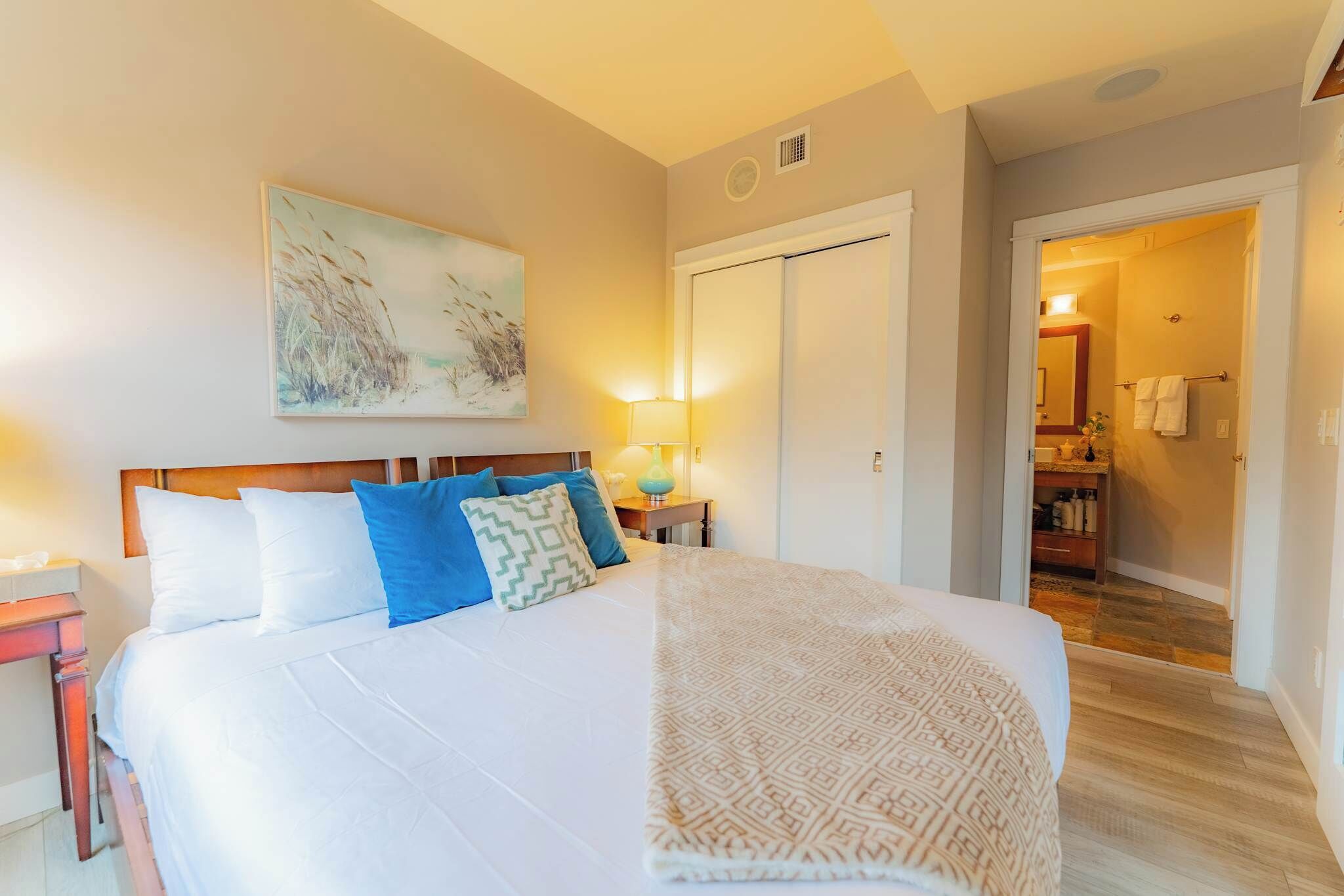 A bedroom with a bed, pillows, a nightstand, a lamp, a painting, and a door leading to a bathroom.