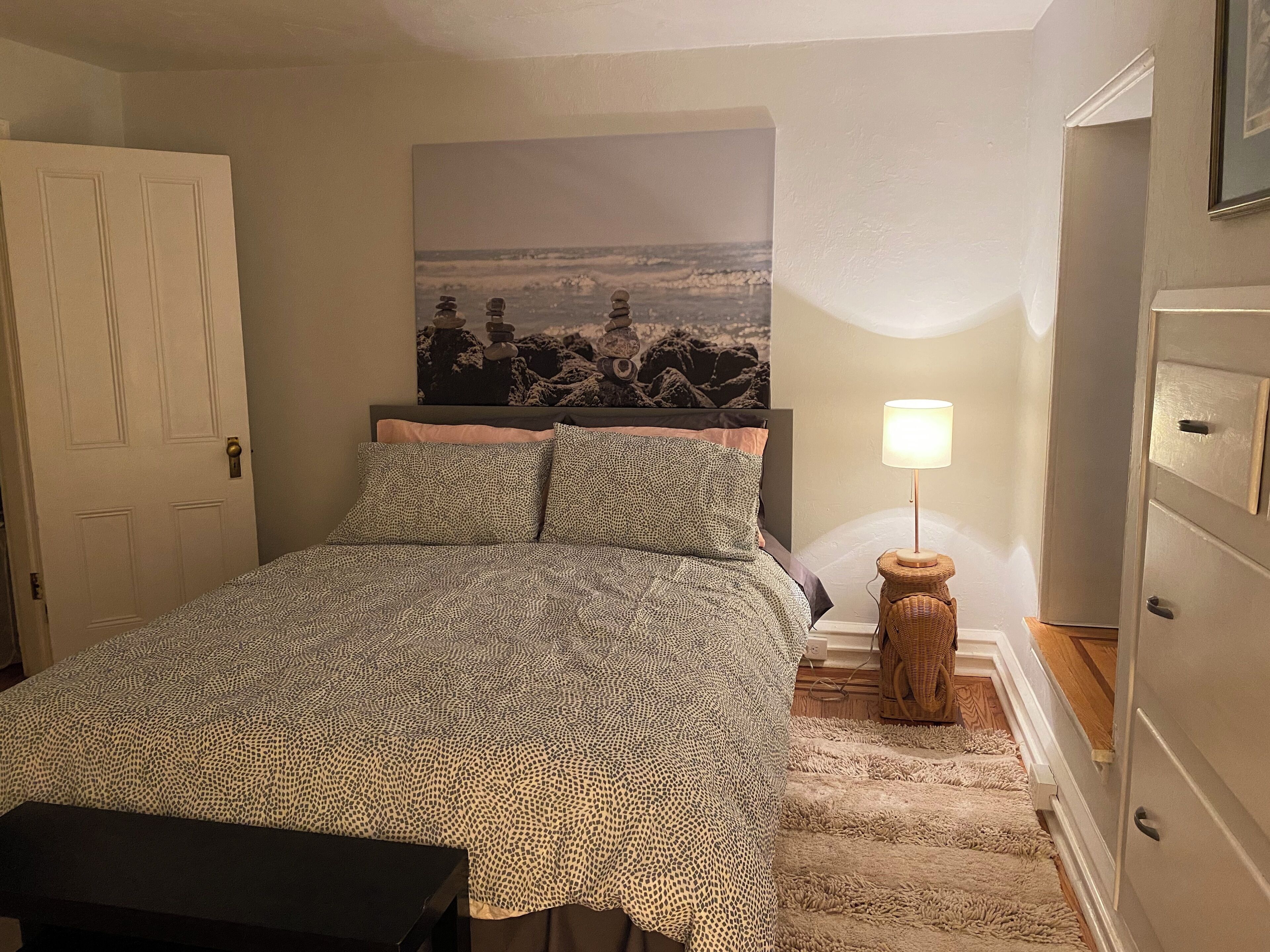 A bedroom with a bed, a nightstand, a lamp, a door, and a wall with a painting.