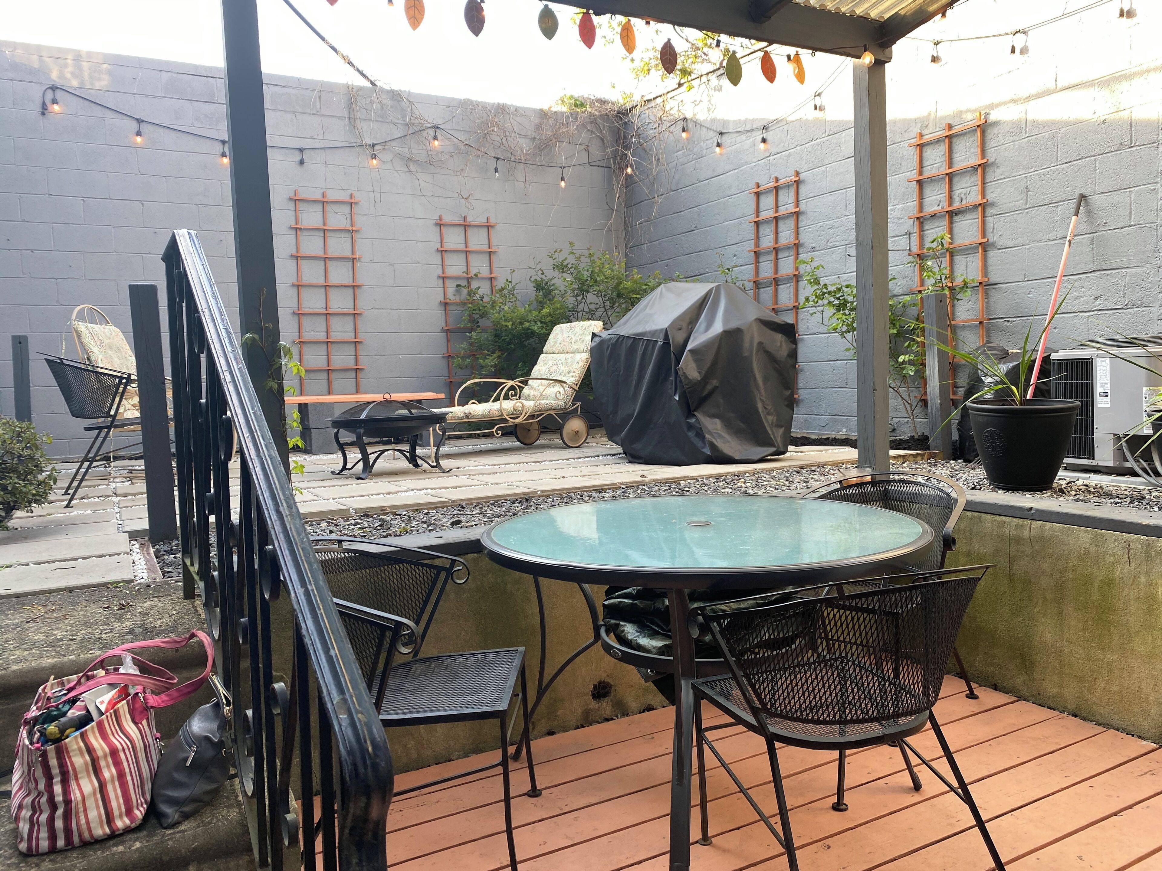 An outdoor patio with a round table and chairs, a covered grill, and a ladder against a grey wall.