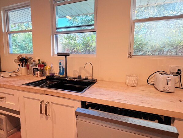 Une cuisine avec un évier noir, des placards blancs et un lave-vaisselle. Des fenêtres offrent une vue sur la verdure environnante.