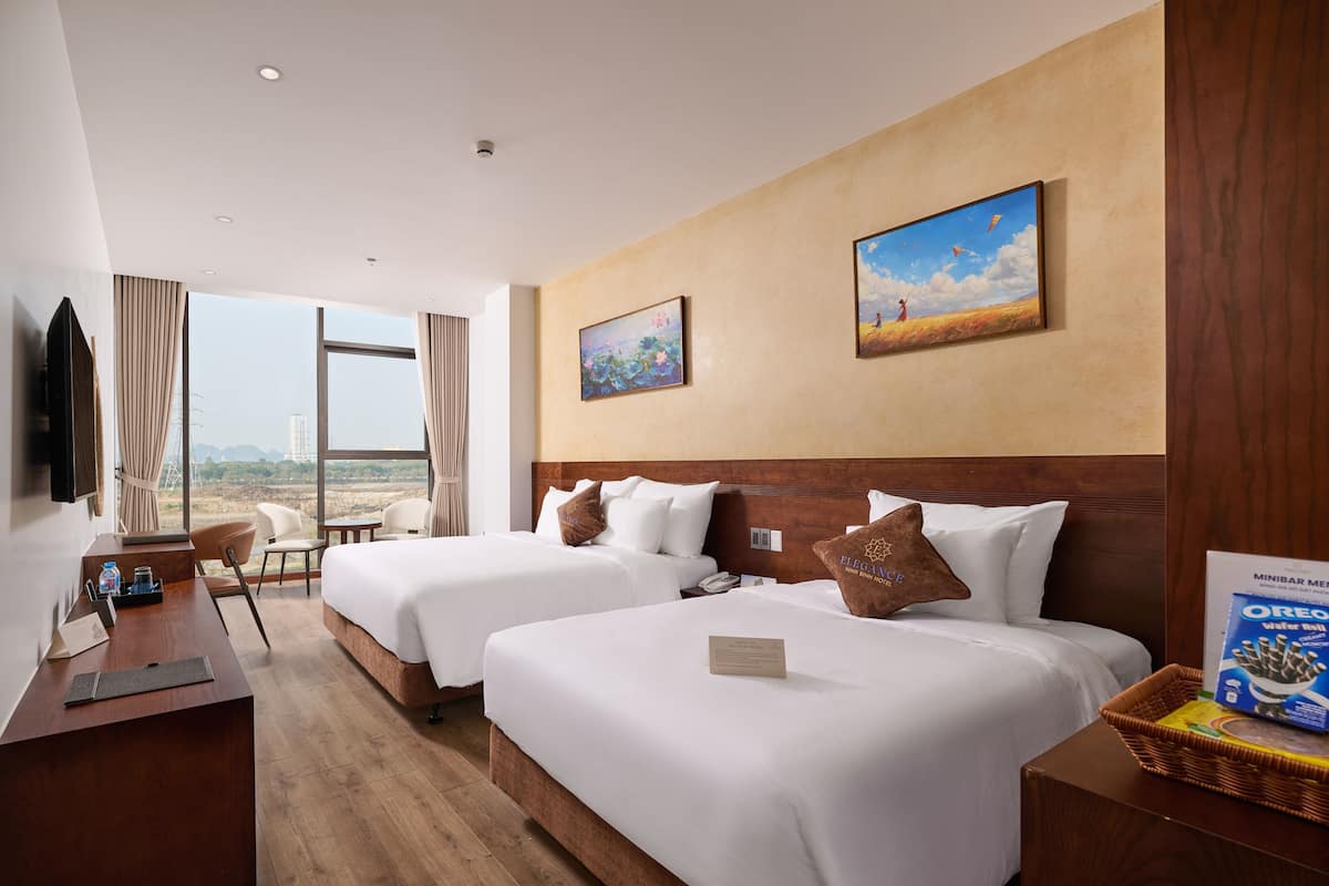A hotel room with two beds, a desk, a chair, a TV, and a large window with a view.